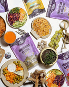A flat lay arrangement of various foods and snacks is displayed on a white surface, including bowls of colorful salads, crackers, grapes, and a drink.
